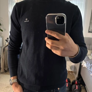 Mörkblå stickad tröja från GANT - Snygg mörkblå stickad tröja från GANT med diskret logga broderad på bröstet. Tröjan har rund halsringning, ribbade muddar och är långärmad. Perfekt för dig som gillar klassisk och stilren look med en modern touch.