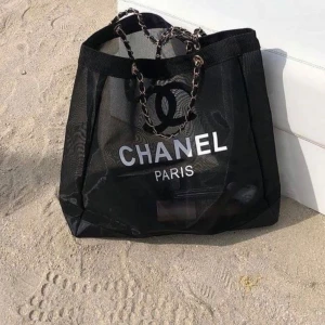 Strandväska - Stor svart strandväska i mesh med tydligt vitt logotryck och text. Väskan har kedjehandtag med inslag av svart material. Perfekt för stranden eller cityhäng, riktigt trendig och rymlig.