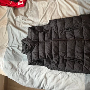 Svart dunväst från Indicode - Säljer en svart dunväst från Indicode med quiltad design och dragkedja framtill. Västen har ståkrage och två fickor på sidorna. Perfekt att slänga över en hoodie för en chill look.