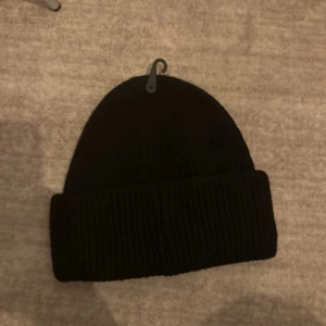 Svart ribbstickad mössa från 157 - En klassisk svart ribbstickad mössa från 157. Mössan har en bred uppvikt kant och är tillverkad i ett mjukt och värmande material. Perfekt för kalla dagar och enkel att matcha med alla outfits.