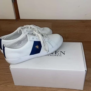 Vita sneakers från Ralph Lauren - Snygga vita sneakers från Ralph Lauren med marinblå detalj och guldfärgad RL-logga på sidan. Skorna har klassisk snörning och är tillverkade i slätt syntetmaterial. Perfekta för en clean och sportig look.