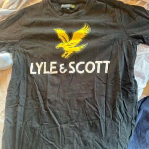 Svart Lyle & Scott t-shirt med tryck - Svart t-shirt från Lyle & Scott med stort gult örntryck och vit logga på bröstet. Klassisk rund halsringning och korta ärmar. Perfekt för dig som gillar streetwear och vill sticka ut med en ikonisk logga.