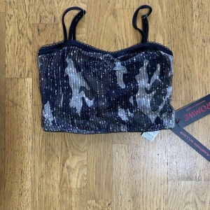 Glittrig camouflage topp från Romwe - Säljer en glittrig croppad topp från Romwe med camouflage-mönster i svart, grått och silver. Toppen har smala axelband och är dekorerad med paljetter över hela framsidan. Perfekt för dig som vill sticka ut med en edgy vibe.
