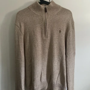 Beige stickad half zip-tröja Polo Ralph Lauren - Beige stickad tröja från Polo Ralph Lauren med ribbad hög krage och half zip-dragkedja. Klassisk broderad logga på bröstet och långa ärmar. Perfekt för lager-på-lager och chill höst- eller vinterstil.