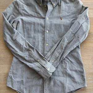 Grå skjorta från Ralph Lauren S - Klassisk grå skjorta från Ralph Lauren i custom fit med button down-krage och broderad logga på bröstet. Skjortan har lång ärm, vita knappar och är tillverkad i bomull. Perfekt för dig som gillar stilren och tidlös look.