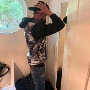 Jordan camo pufferjacka XL - Snygg pufferjacka från Jordan i camomönster med grå, beige och bruna toner. Jackan har svarta ärmar med röd JORDAN-text, röd dragkedja och huva med röd insida. Perfekt för dig som vill sticka ut med streetstyle-vibe.