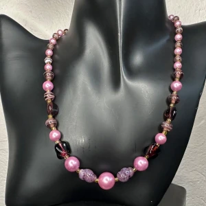 Färgstarkt vintagehalsband med rosa & lila pärlor – Retrofynd från 60-/70-talet - Vintagehalsband med pärlor i rosa, lila och vinrött – en lekfull blandning av glas, mönstrade och lackade pärlor. Inropat på auktion med äldre silversmycken och bijouterier. Ca 40 cm långt, största pärlor ca 1 cm. Sannolikt europeiskt och från sent 60-tal till tidigt 80-tal. I fint vintageskick. Ingen prutning, men jag erbjuder rabatt vid köp av flera saker. Passa på – kolla gärna mina andra annonser också!  