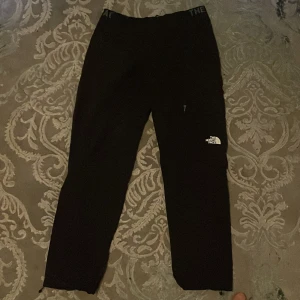 Svarta täckbyxor från The North Face - Svarta täckbyxor från The North Face med elastisk midja och logga på benet. Byxorna har dragkedja vid fickan och är perfekta för kalla dagar. Materialet är slitstarkt och passar för utomhusaktiviteter. Snygg och sportig look med normal passform. Byxan är gjort av nylon och polyester.