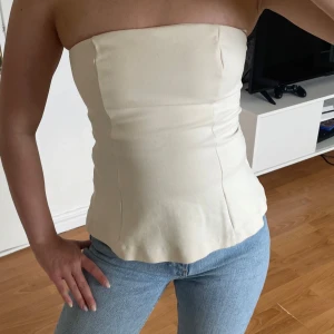  bandeau topp - Säljer en stilren krämvit bandeau tubtopp med figurnära passform. Toppen är axelbandslös och har en lätt utsvängd nederkant. Perfekt att matcha med jeans eller kjol för en clean look. Aldrig använd 😊