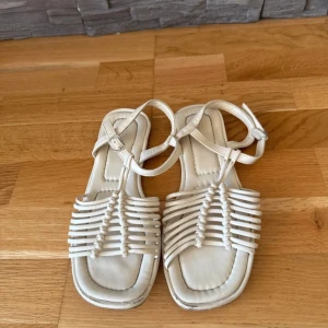 Vita sandaler med flätade remmar - Snygga vita sandaler med flätade remmar och fyrkantig tå. De har justerbart spänne runt ankeln och en platt sula för skön komfort. Perfekta för varma dagar och ger en clean look till din outfit. Beige, Crémefärgad