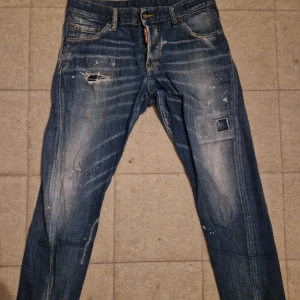 Blå Dsquared2 jeans med slitningar - Säljer ett par blå jeans från Dsquared2 med coola slitningar, patch och färgstänk. Jeansen har klassisk femficksdesign, raka ben och tydliga slitdetaljer framtill. Märkespatch på bakfickan och snyggt tvättad denim ger en edgy vibe. Prisen kan diskutera dm