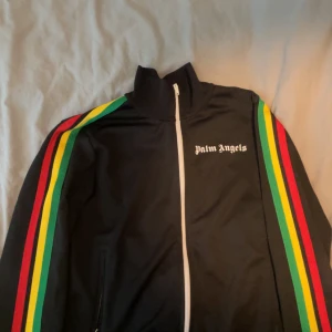 Palm Angels svart vindjacka med ränder - Svart vindjacka från Palm Angels med vita dragkedjor och färgglada ränder i rött, gult och grönt längs ärmarna. Jackan har hög krage och logga på bröstet. Perfekt för en sportig och trendig look. 