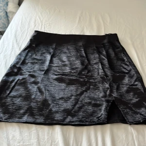Svart satin-kjol från Gina Tricot - Supersnygg svart kortkjol i glansigt satinmaterial från Gina Tricot. Kjolen har en liten slits framtill som ger en extra edge. Perfekt för dig som gillar stilrena och trendiga plagg med en lyxig känsla.