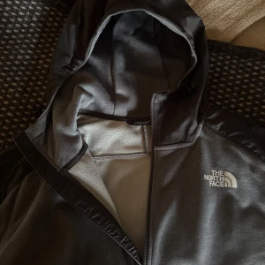Grå fleece-jacka från The North Face - Riktigt schysst grå fleece-jacka från The North Face med mörkgrå huva och axelparti. Jackan har dragkedja hela vägen, två fickor och en stilren design. Perfekt till både träningstillfällen, utomhus men även perfekt till vardagliga outfits. Insidan är full fleece, skydar mycket väl mot vind och håller sig varm samtidigt som den är väldigt lätt och tunn. Den är använd bara 1-2 gånger, alltså nästan nyskick. Ordinarie pris 1300