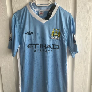 Manchester City Aguero tröja Umbro - Manchester Citys ljusblå matchtröja med vita detaljer, Umbro-logga och klubbmärke på bröstet. Tryck med Etihad Airways på framsidan och Kun Agüero 16 på ryggen. Klassisk V-ringning och korta ärmar. Perfekt för dig som älskar fotboll och city vibes.
