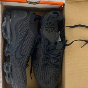 Nike Vapormax svarta sneakers strl 42,5 - Svarta Nike Vapormax sneakers med stickad ovandel och genomskinlig bubblig sula. Snyggt diskret mönster med små färgprickar i tyget och klassisk svart swoosh på sidan. Skorna har snörning och är riktigt lätta och flexibla – perfekta för dig som gillar streetstyle. och aldrig har använt Helt nya för ett liten summa.