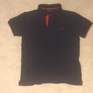 Svart pikétröja från Gant med röda detaljer - Svart pikétröja från Gant med röda detaljer vid krage, knäppning och ärmslut. Klassisk passform och kortärmad modell med broderad Gant-logga på bröstet. Perfekt för en stilren och sportig look.