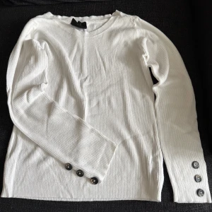 Vit ribbad långärmad tröja med knappar - Vit ribbad långärmad tröja med rund hals och snygga svarta knappar på ärmsluten. Tröjan har en clean och stilren look som passar perfekt till jeans eller kjol. Materialet känns mjukt och stretchigt, vilket gör den riktigt skön att ha på sig.