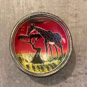 Skål - Bilderna visar en handmålad skål med motiv av en giraff som äter från ett träd mot en färgstark bakgrund. Skålen har konstnärliga detaljer och passar perfekt som dekoration eller förvaring av småsaker. 