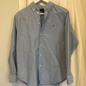 Ljusblå skjorta från Ralph Lauren - Helt ny Klassisk ljusblå skjorta från Ralph Lauren med button-down krage och broderad logga på bröstet. Skjortan har långa ärmar, knappar framtill och är tillverkad i mjuk bomull. Perfekt för en clean och stilren look.
