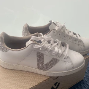 Vita Victoria sneakers med glitterdetaljer - Snygga vita sneakers från Victoria med glittriga silverdetaljer på hälen och sidan. Klassisk rund tå, vita snören och platt sula. Perfekta för dig som vill ha en stilren sko med lite extra bling. Tillverkade i syntetmaterial.