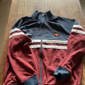 Ellesse vinröd och marinblå trackjacka XL - Snygg trackjacka från Ellesse i vinrött och marinblått med vita ränder över bröstet och ärmarna. Jackan har dragkedja hela vägen, höga muddar och klassisk sportig look. Perfekt för dig som gillar retrostil och streetwear.