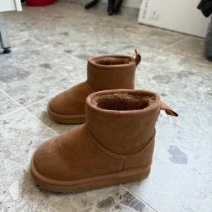 Bruna boots från H&M, storlek 26/27 - Mysiga bruna boots från H&M med mjukt foder och platt sula. Skorna är i mockaliknande material och har en rund tå samt dragflik baktill. Perfekta för kyliga dagar när du vill hålla fötterna varma och sköna.
