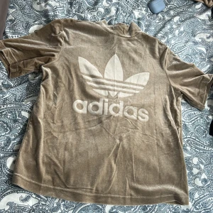 Beige sammetst-shirt från Adidas - Säljer en beige t-shirt i sammet från Adidas med stort vitt Trefoil-logga på ryggen. Färgen är lite ljusare än vad det ser ut på första bilden