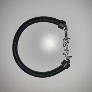 YSL svart armband med silverdetaljer - Snyggt svart armband från YSL med grovt band i skinn och coola silverdetaljer. Armbandet har ett ikoniskt YSL-hänge i glittrande silver och dekorativa metalldetaljer vid fästena. Perfekt accessoar för att lyfta din outfit.
