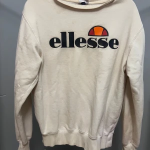 Vit sweatshirt från Ellesse - Säljer en vit sweatshirt från Ellesse med stor logga framtill i svart, orange och rött. Tröjan har rund hals, långa ärmar och ribbade muddar vid ärmslut och nederkant. Perfekt för en chill och sportig look.