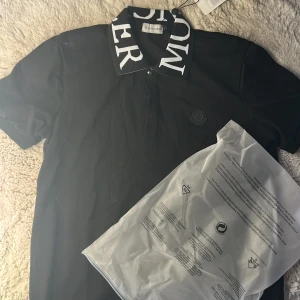 Svart Moncler t-shirt med logga - Svart t-shirt från Moncler med korta ärmar och stor vit logotyptext på kragen. Klassisk pikékrage och diskret Moncler-märke på bröstet. Snygg och stilren modell i bomull, perfekt för dig som gillar exklusiva streetwear-vibbar.