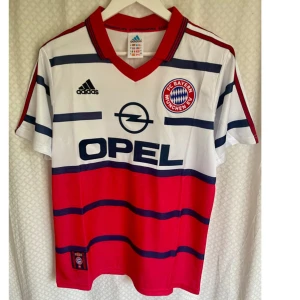 Vintage Jersey FC Bayern München  - Vintage Fc Bayern München match hemmatröja säsong 1998/1999 från Adidas med klassiskt Opel-tryck, röd och vit bas med mörkblå horisontella ränder. Nummer 10 och Matthäus på ryggen. Röd krage och tre ränder på axlarna. Perfekt för samlare eller fans av 90-talets fotboll.