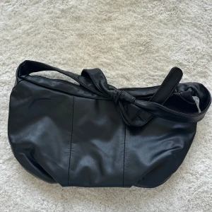 Svart axelväska med knytband   - Snygg svart axelväska i skinnimitation med mjuk, rundad form och ett brett axelband som knyts i en cool knut. Väskan har dragkedja upptill och är perfekt för att få plats med allt du behöver. Enkel och stilren design som funkar till många outfits.