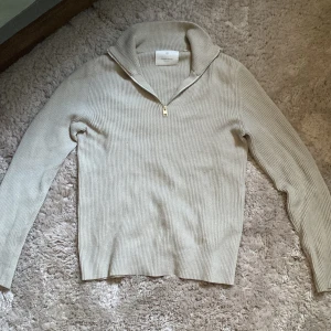 Beige ribbad stickad tröja Jack & Jones - Säljer en beige stickad tröja från Jack & Jones med ribbad struktur och half zip-dragkedja vid halsen. Tröjan har långärmade och en snygg krage som går att vika ner. Perfekt för dig som gillar stilrena och enkla plagg med lite edge.