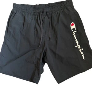Svarta Champion badbyxor - Svarta shorts från Champion med vit och röd logga längs benet. De har elastisk midja med snörning, sidofickor och en bakficka med lock. Meshfoder på insidan för extra komfort. Perfekta för chill eller sportiga dagar.