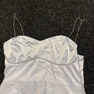 Ljusblå långklänning med strassband - Säljer en elegant ljusblå långklänning med glansigt satinmaterial och tunna axelband dekorerade med strass. Klänningen har en v-ringad byst och ett strassband som följer bystlinjen för extra bling. Perfekt för dig som vill sticka ut med en stilren look.