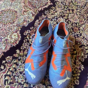 Puma Future Elite - Säljer ett par snygga fotbollsskor från Puma i blått och orange med vita Puma-loggor. Skorna har snörning och en platt sula med dobbar för gräsplan. Ovandelen är i syntetmaterial och designen är riktigt sportig och modern. Dom är använf i en match och inte mera efter det på grund av att de va för små