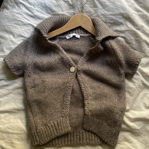 Beige stickad croppad kofta från Zara - Säljer en beige stickad kofta från Zara med korta ärmar och stor krage. Koftan är croppad och har en knapp framtill. Perfekt att styla med jeans eller kjol för en chill look. Materialet är mjukt och ribbat vid muddar och krage.