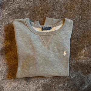 Grå sweatshirt från Polo Ralph Lauren - Snygg grå sweatshirt från Polo Ralph Lauren med klassisk rund hals och broderad vit logga på bröstet. Tröjan har långa ärmar, ribbade muddar och är gjord i mjuk bomullsmix som är skön mot huden. Perfekt för chill dagar och enkel att matcha med jeans eller joggers. Passar folk som har storlek S.