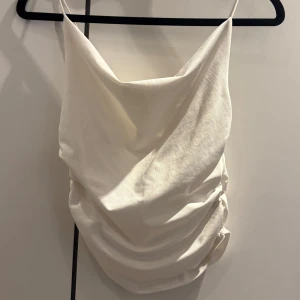 Vit draperad linnetopp med tunna band - Säljer en snygg vit linnetopp med tunna axelband och draperad framsida. Toppen har en lätt glansig yta och rynkade detaljer i sidorna som ger en cool och trendig look. Perfekt att styla med jeans eller kjol för en fräsch vibe.