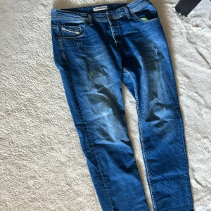 Blå Diesel jeans herr - Snygga blå Dieswl jeans med klassisk femficksdesign och coola slitningar framtill. Jeansen har en regular och raka ben, med detaljerad logotyp på bakfickan och läderpatch i midjan. Perfekt för dig som gillar streetstyle och vill ha ett par jeans med attityd.
