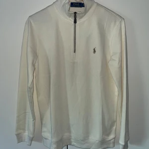 Vit half zip tröja från Polo Ralph Lauren - Snygg vit långärmad tröja från Polo Ralph Lauren med half zip och klassisk broderad logga på bröstet. Tröjan har en stilren design med hög krage och är gjord i mjuk bomull som känns skön mot huden. Perfekt för dig som gillar clean och enkel stil.