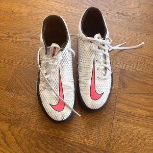Nike Phantom fotbollsskor vit/rosa - Nike Phantom fotbollsskor i vit med svarta och rosa detaljer. Snyggt mönstrad ovandel med stor rosa Swoosh på sidan och snörning framtill. Platt sula i svart och vit, designad för inomhusspel. Skorna är tillverkade i syntetmaterial och har en sportig look. Ingen låda tyvärr