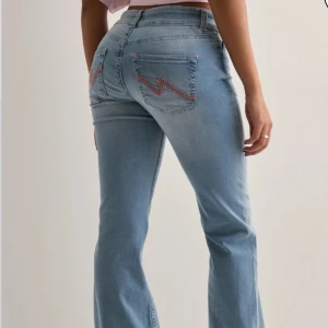 Bootcut ljusblå jeans - Snygga ljusblå lågmidjade bootcut jeans med två knappar fram och broderad detalj i rosa på bakfickorna. Jeansen har klassisk femficksdesign och är tillverkade i mjukt denimtyg som sitter bekvämt på kroppen. Perfekta för dig som gillar en chill och trendig look.