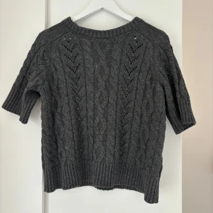 Mörkgrå stickad tröja från Zara - Säljer en mörkgrå stickad tröja från Zara med korta ärmar och snyggt kabelstickat mönster. Tröjan har bred ribbad kant vid hals, ärmslut och nederkant. Perfekt för lager-på-lager och chill höststilar.