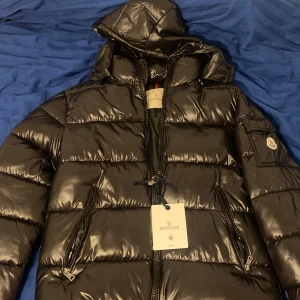 Svart Moncler dunjacka med huva - Säljer en svart, glansig dunjacka från Moncler med huva och dragkedjor på fickorna. Jackan har ett klassiskt Moncler-märke på ärmen och quiltad design. Perfekt för kalla dagar och har en snygg, modern look som passar streetstyle.