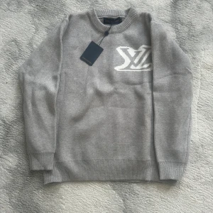 Grå stickad Louis Vuitton tröja - Snygg grå stickad tröja från Louis Vuitton med stort vitt LV-logo på bröstet. Rund hals och långa ärmar med ribbade muddar. Perfekt för dig som gillar exklusiva märken och stilren design. Materialet är mjukt och skönt mot huden. Pris kan diskuteras vid snabb affär.