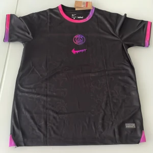 Svart PSG fotbollströja Nike L - Snygg svart PSG fotbollströja från Nike med Dri-FIT material som andas. Tröjan har korta ärmar och coola detaljer i rosa och lila på krage och ärmslut. Framsidan har PSG-logga och ett unikt rosa Nike-märke, baksidan har namnet Rikardo och nummer 77 i rosa och lila.