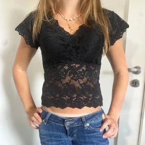 Svart spetsblus med v-ringning - Supersnygg svart blus med v-ringad hals och korta ärmar. Blusen är croppad och har ett genombrutet spetsmönster med blommiga detaljer. Perfekt att styla med jeans eller kjol för en trendig look.