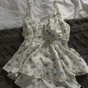 Blommig vit kortklänning med rosett - Supersöt vit kortklänning med blått blommönster och smala axelband. Klänningen har knappar framtill, markerad midja och en stor rosett i ryggen. Den är lätt och luftig med volangdetaljer, perfekt för varma dagar. Materialet känns som tunn polyester eller bomull.